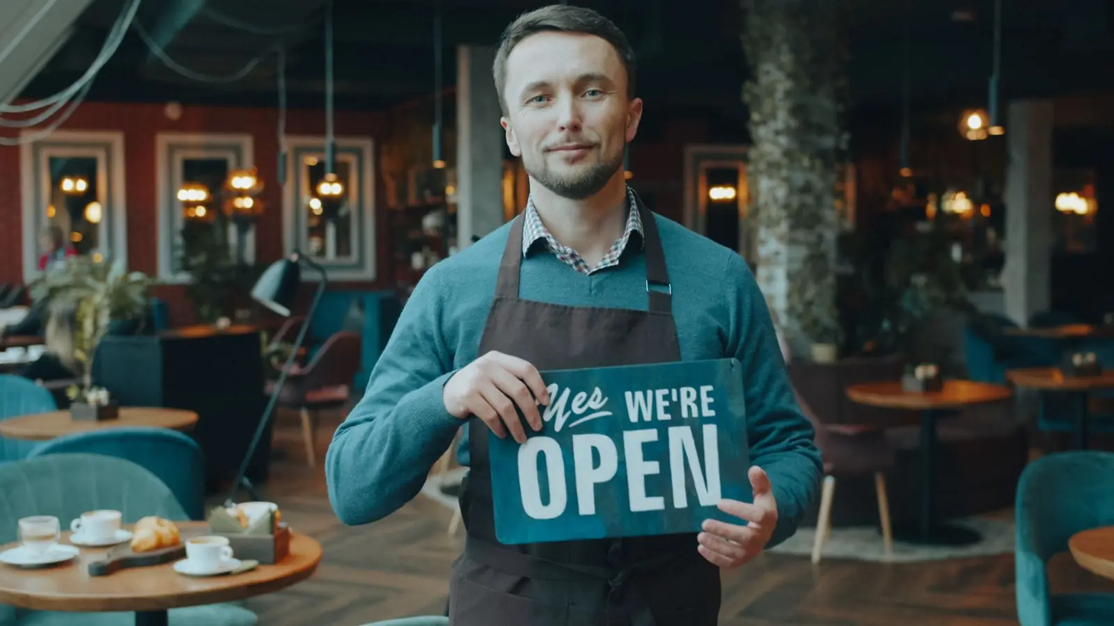 Cafe owner standing inside, holding a 'We're Open' sign, welcoming customers.