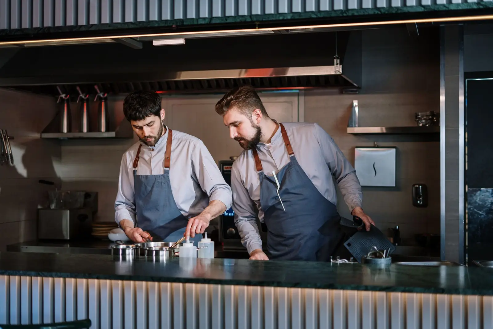 Two male chefs in aprons cook attentively in a contemporary open kitchen setting.